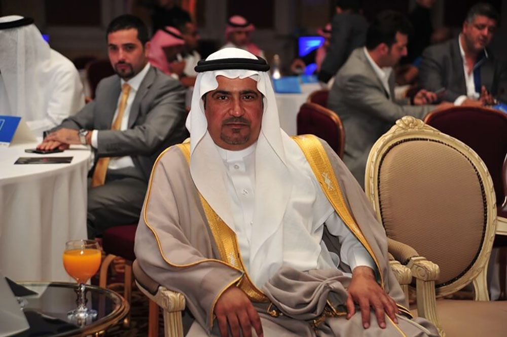 Man in traditional Arab dress, seated, looking at camera, in a formal setting. Orange juice on table.