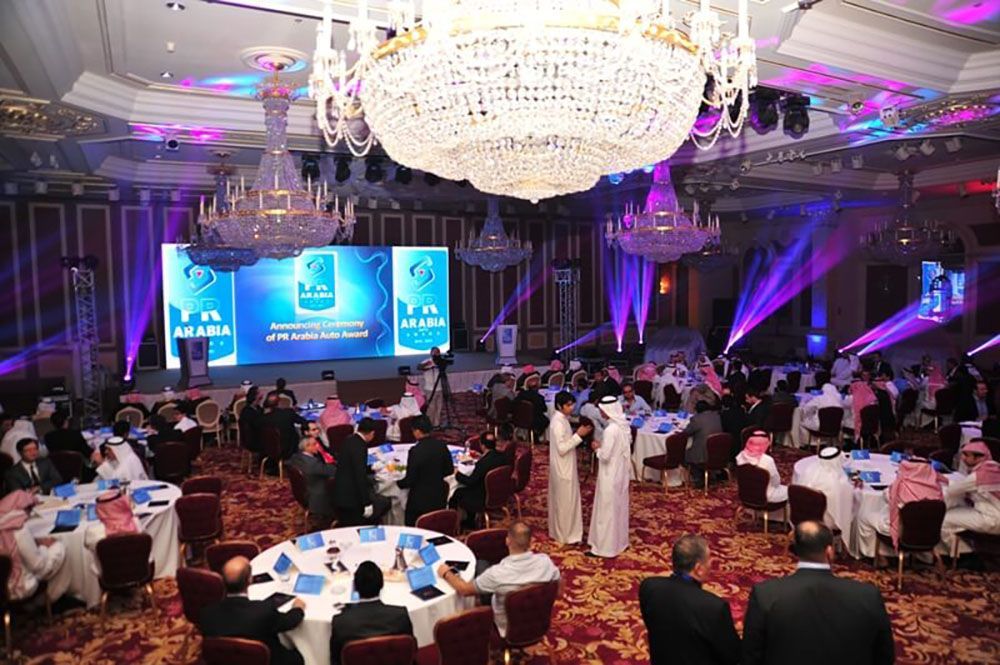 An event at a ballroom with people at tables, a stage, and chandeliers.