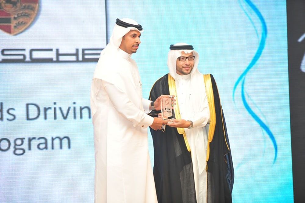 Two men in traditional Arab dress, one presenting an award. Porsche logo in background.