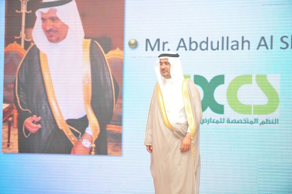 Man in traditional Arab attire stands next to a screen featuring his image and name.