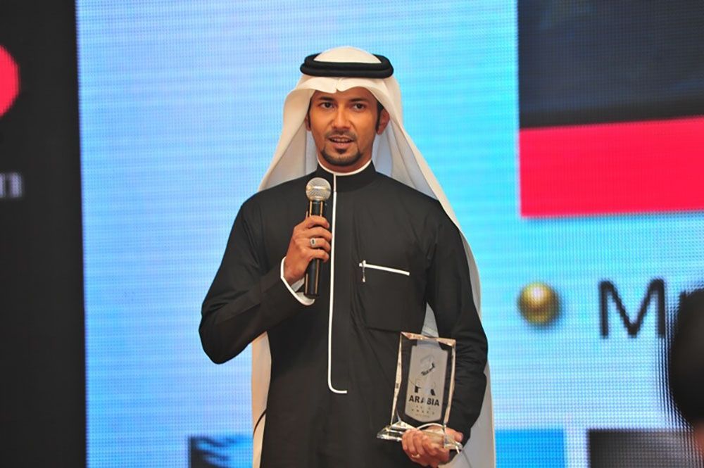 Man in traditional attire holding a microphone and an award, speaking at an event.