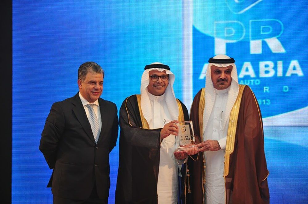 Three men in suits and traditional attire holding an award at an event with a blue backdrop.