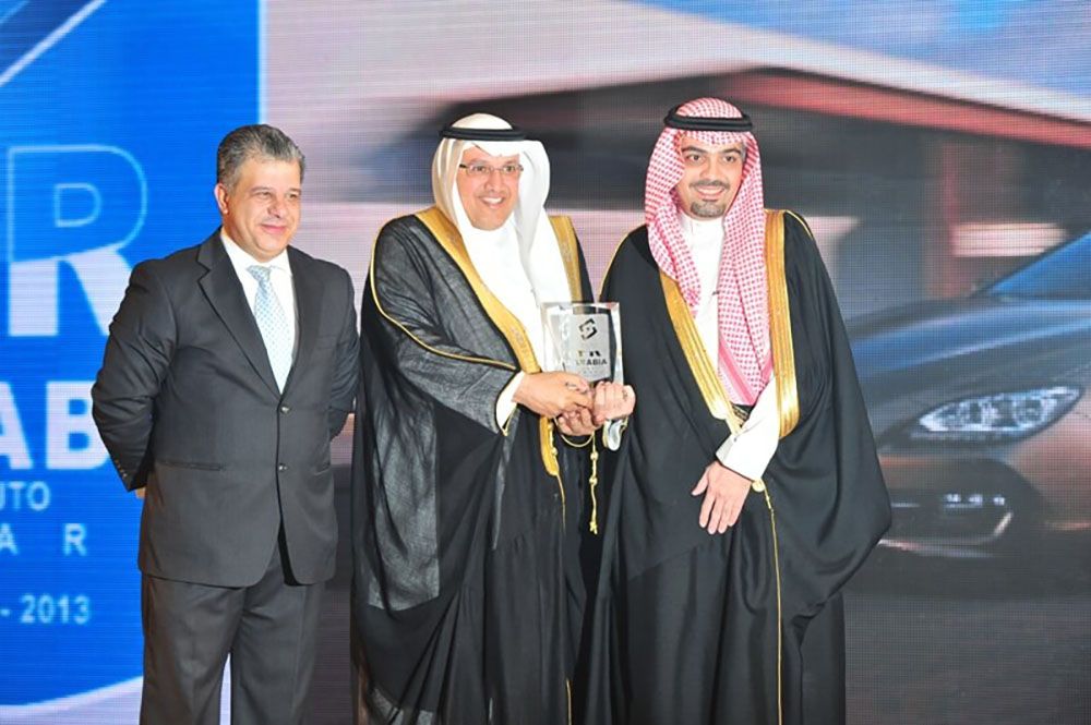 Three men at an auto show. Two in traditional robes, one in a suit, holding an award.