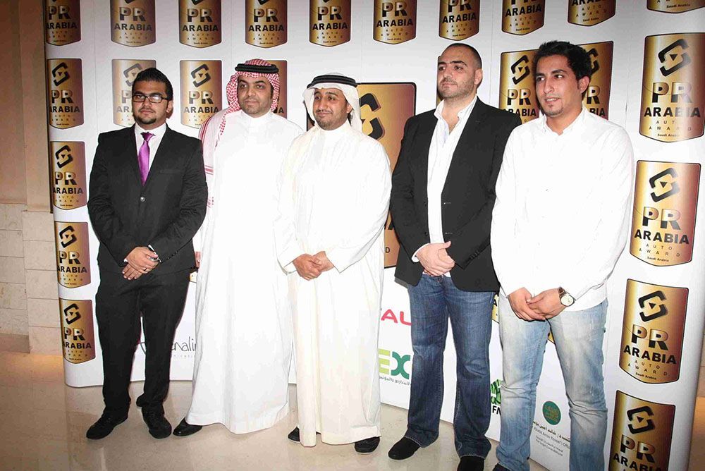Five men posing, some in suits, some in traditional Arab attire, at an event with a branded backdrop.