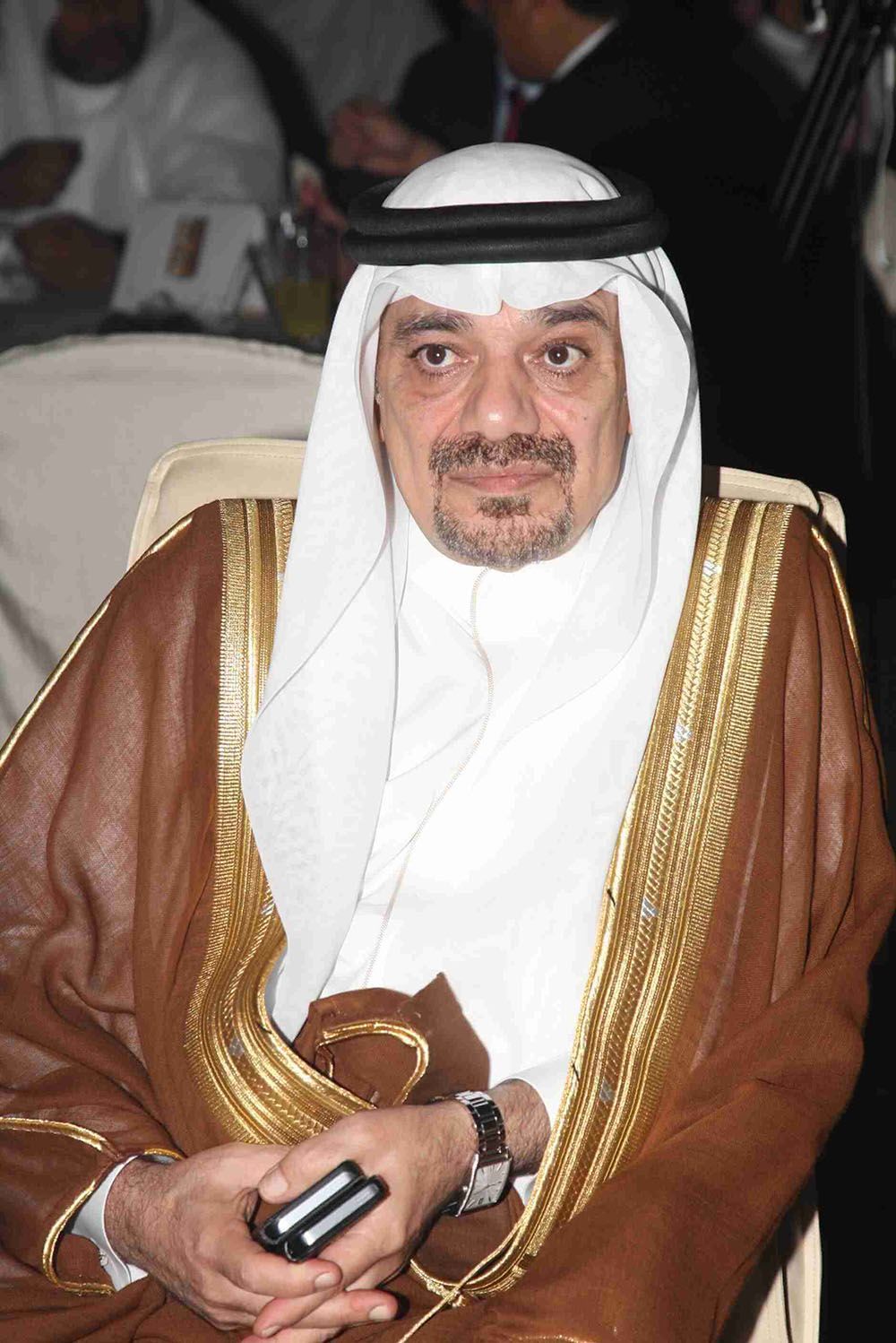 Man in traditional Arabic attire, seated with a neutral expression, holding a phone.