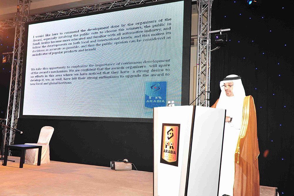 Man in traditional dress speaking at a podium in front of a screen displaying text.