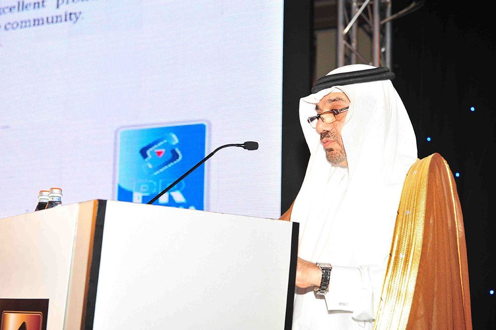 Man in traditional Arab attire speaking at a podium; blue logo on the backdrop.
