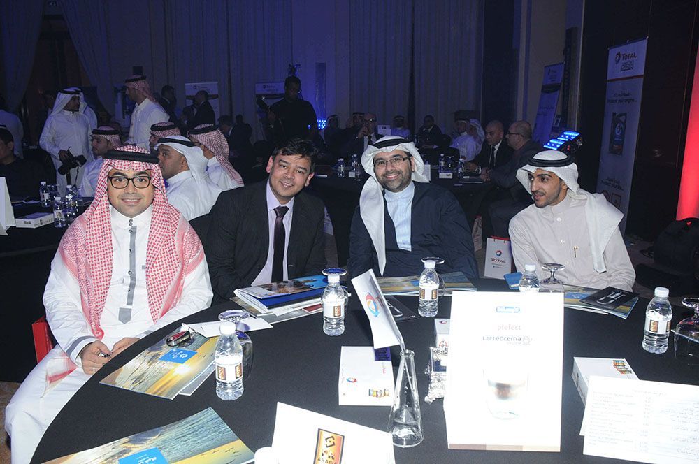 Group of men, some in traditional attire, at a table with promotional materials, inside event.