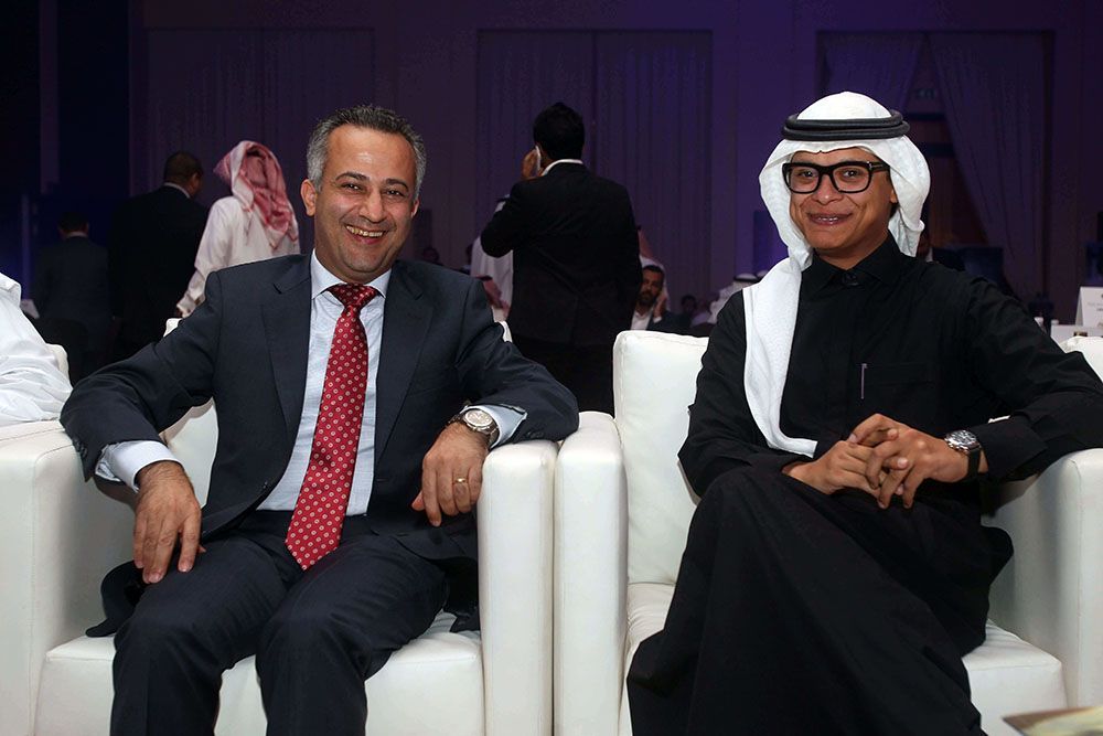 Two men in suits sit on white chairs at an event. One wears a suit and red tie. The other wears a thobe and a shemagh.