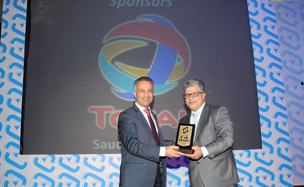 Two men in suits exchanging an award in front of a Total logo backdrop.