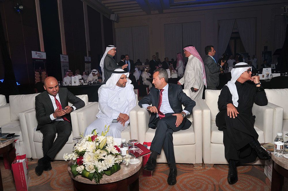 Men in formal attire and traditional thobes at a gathering, seated in a lounge area, conversing.