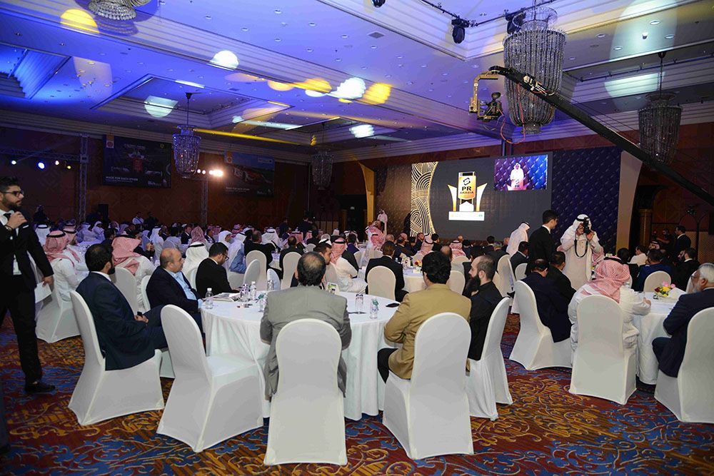 A large, formal event with people seated at round tables, a stage in the background, and decorated ceilings.