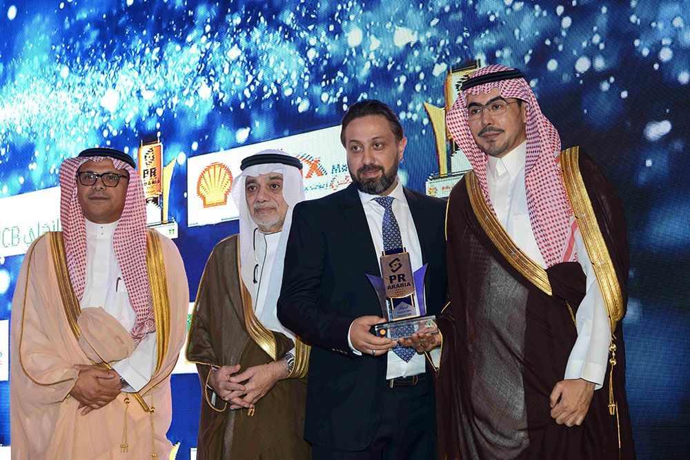 Four men at an award ceremony. A man in a suit holds a trophy, flanked by men in traditional robes, against a stage backdrop.
