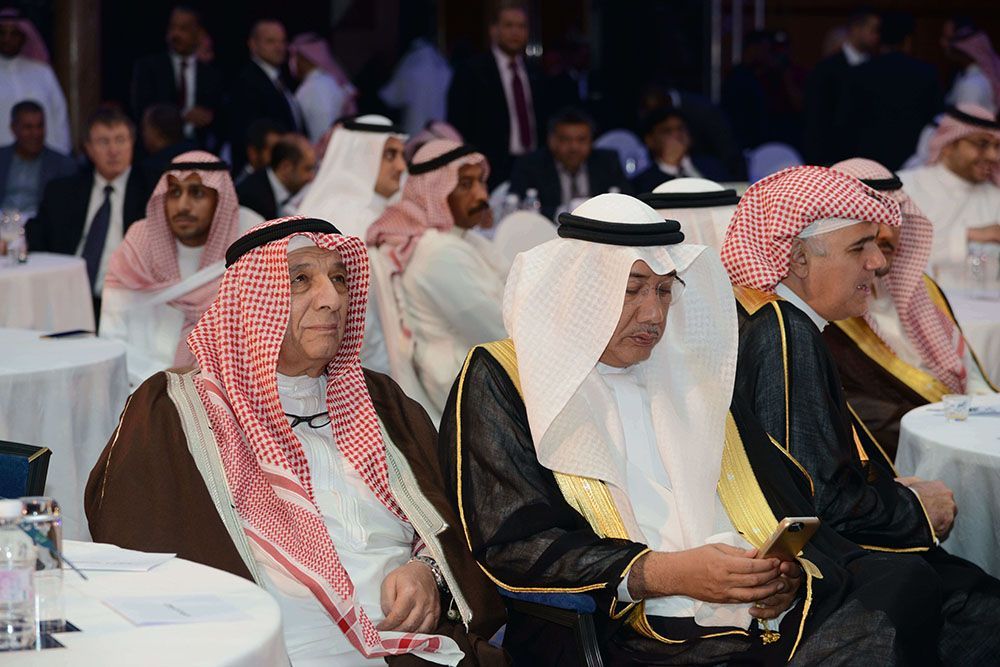 Men in traditional Arab attire at a formal event, some looking at phones, seated at tables.