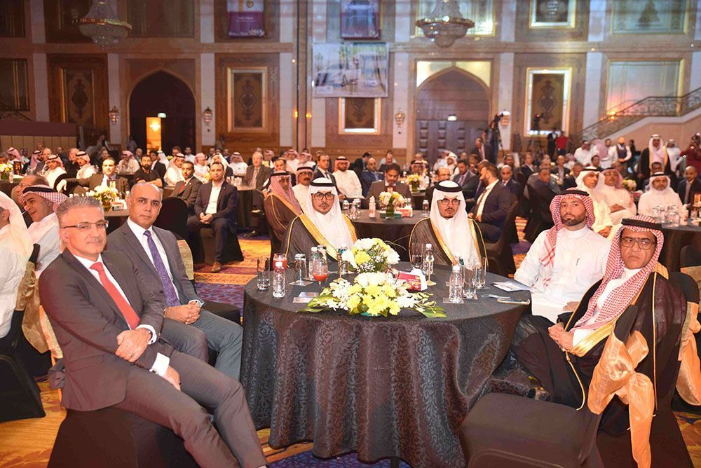 Attendees at an event seated at tables. Some wear traditional Arab attire, others suits. Ornate hall.