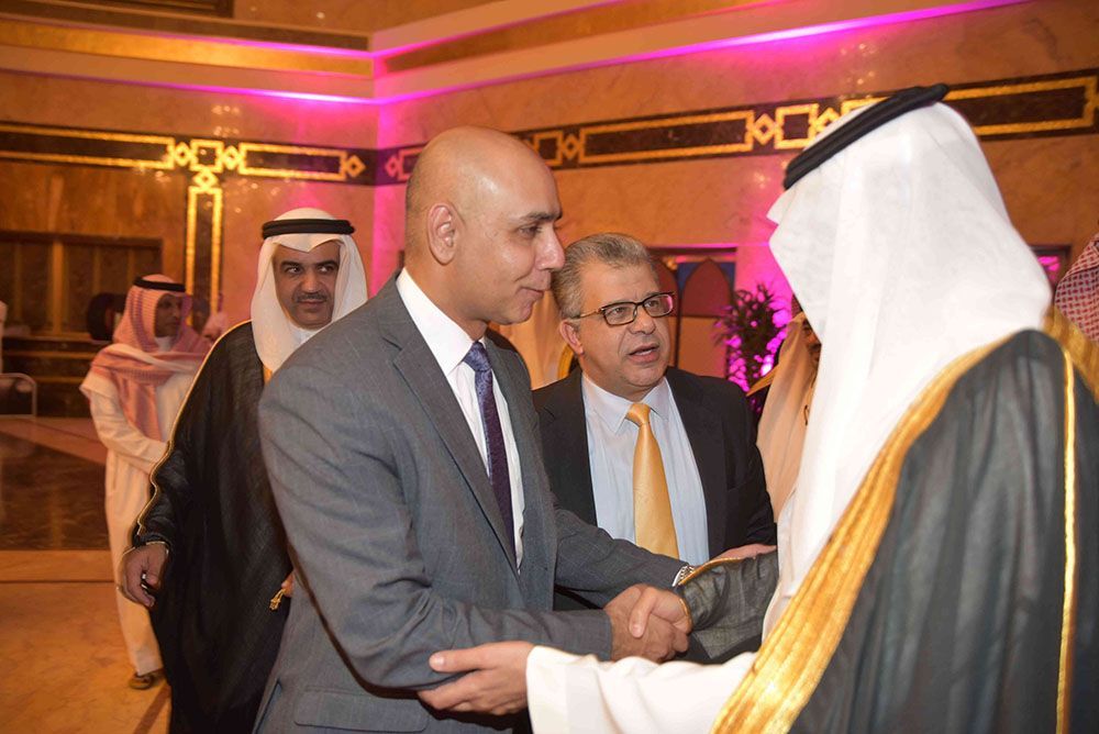 Men in formal attire shake hands at an event. One wears traditional Saudi clothing.