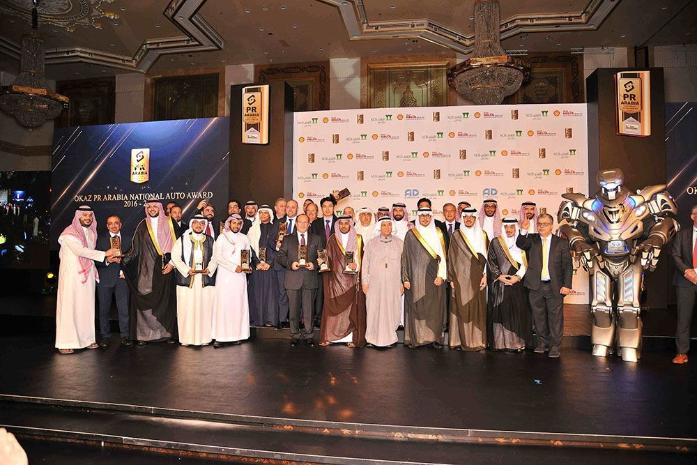 Award ceremony with group of people holding trophies, robot, stage backdrop.