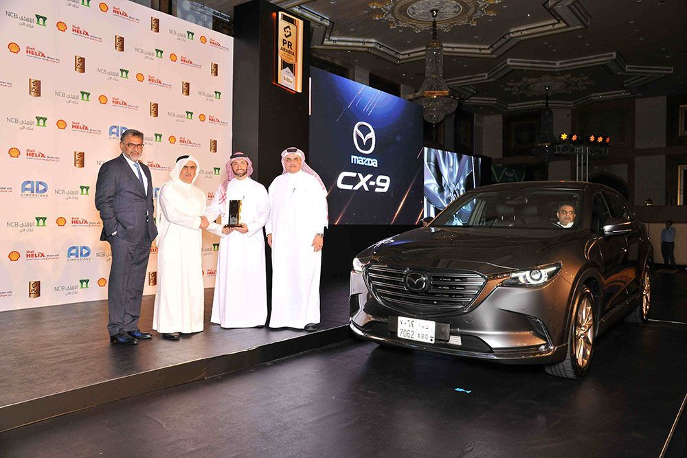 Men presenting a Mazda CX-9 on a stage. Some wear suits and traditional white robes. Dark car with a screen.