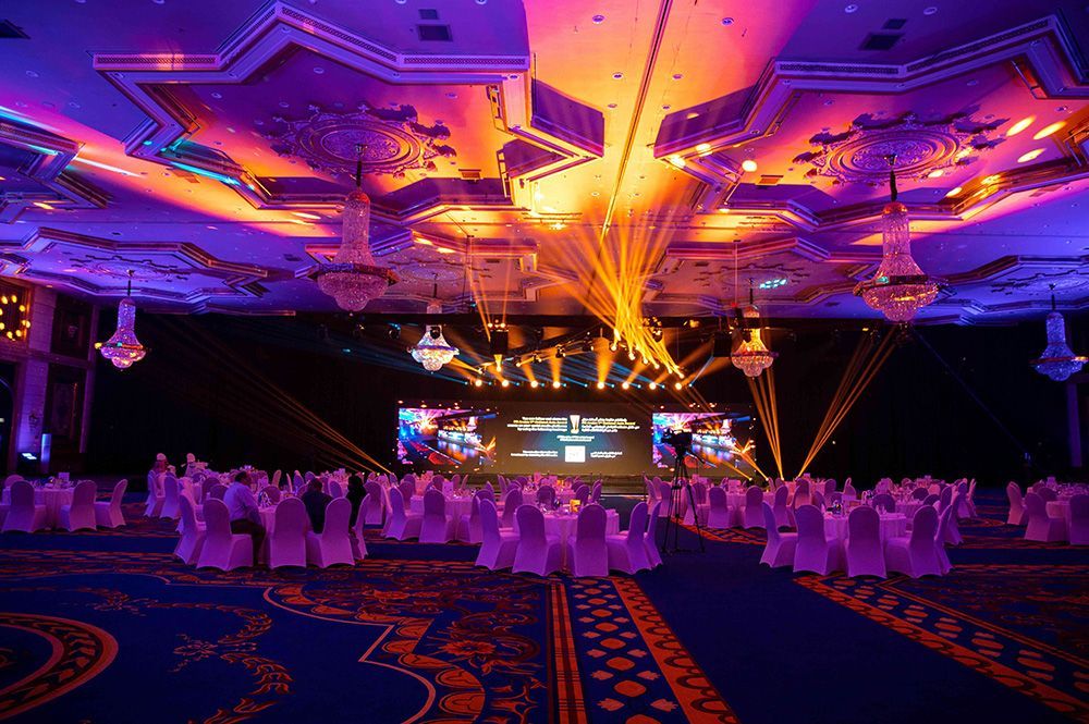 Formal event hall decorated with purple and orange lighting. Rows of tables face a stage with a screen.