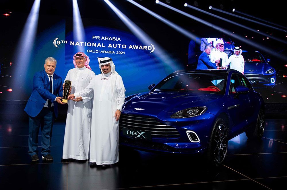 Men in white robes and suit presenting an award beside a blue Aston Martin SUV on a stage.