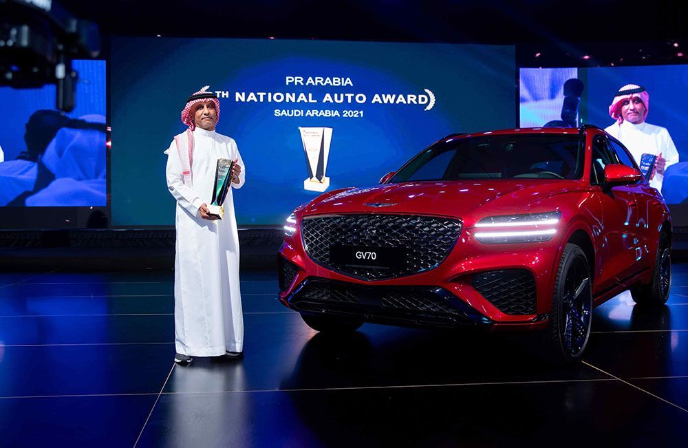 Man in traditional white attire holding an award, standing next to a red Genesis GV70 SUV at an event.
