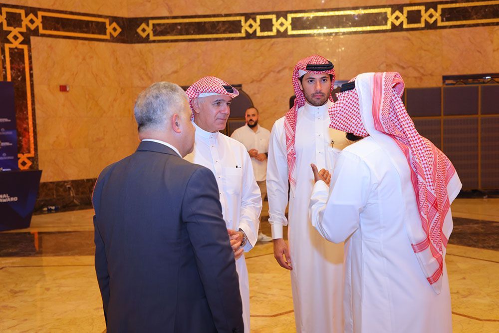 Four men in a room, some in traditional white robes and red/white headscarves, talking. One man in a suit.