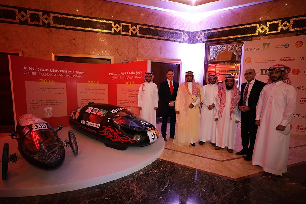 Group of men in traditional attire with two Shell Eco-marathon cars on display in a decorated hall.