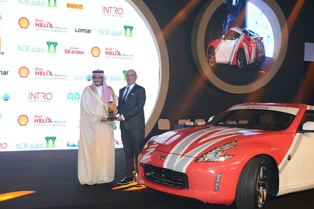 Two men at an award ceremony, one presenting a trophy. Red and white sports car on display.