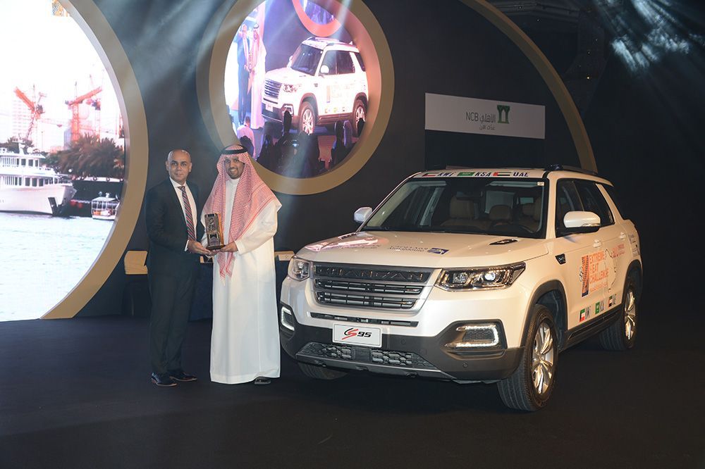 Two men at an event beside a white SUV; one holds an award. Stage setting, white and gold colors.
