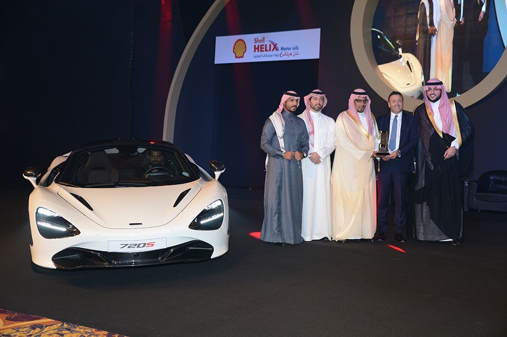 White McLaren sports car with a group of men in formal attire and a Shell Hi-Lix banner.