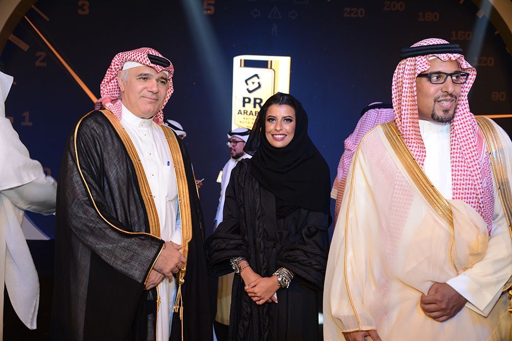 Three people in traditional Arab dress, smiling at an event, standing in front of a logo.