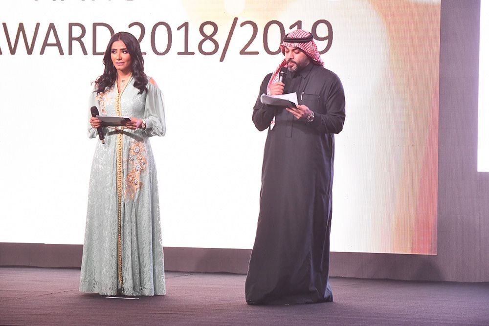 Two people on stage at an event. Woman in blue dress, man in traditional Arab attire.