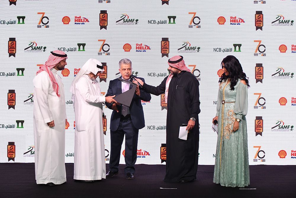 Award ceremony: Men in traditional Arab attire and suits on stage. Woman in gown stands beside them.