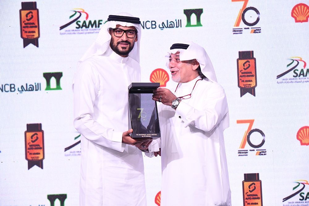 Two men in white thobes exchange a trophy on a stage with logos in Arabic and English.