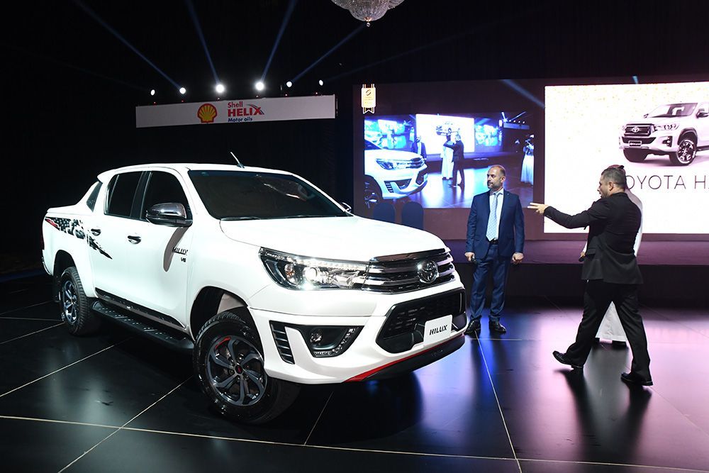 White Toyota Hilux pickup truck at a launch event, two men in suits gesturing.