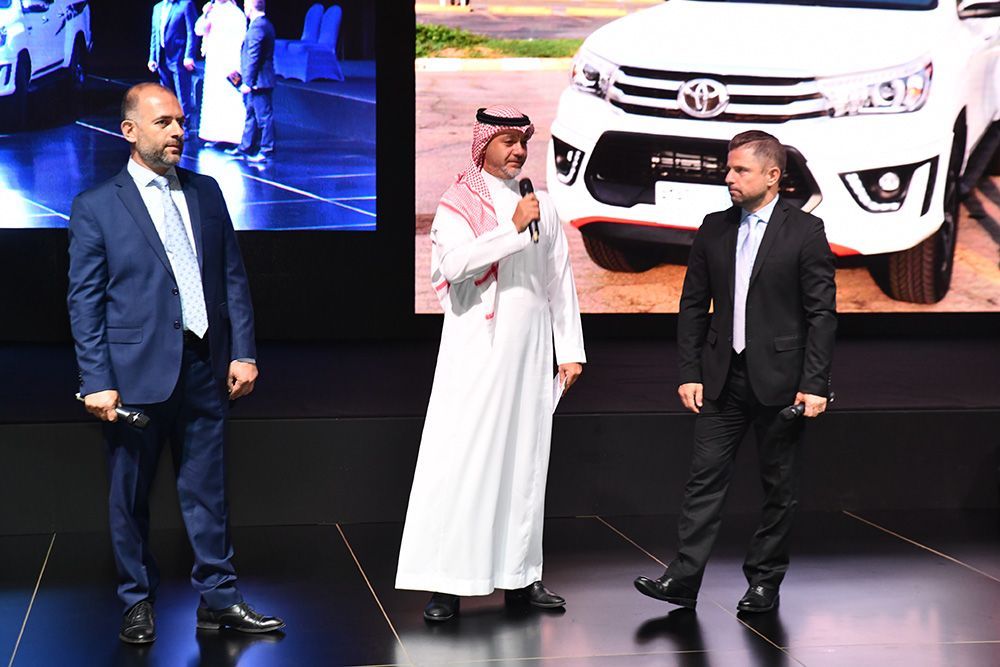 Three men on a stage; one in traditional Arab dress speaking into a microphone, flanked by two men in suits, with a Toyota vehicle in the background.