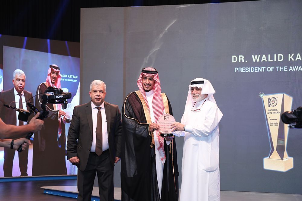Man in traditional attire receives an award. Two men in suits and a cameraman stand by on a stage.