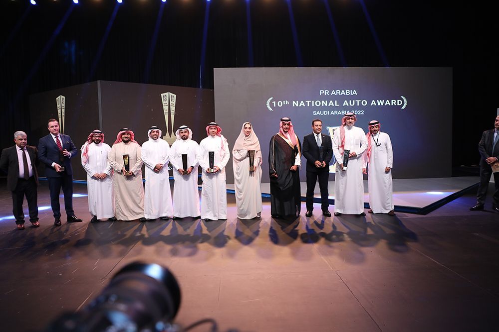 A group of men in formal wear and traditional robes are on a stage, holding awards.
