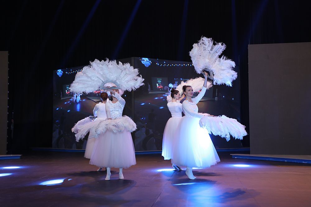 Dancers in white gowns with feathered fans performing on stage, blue lights illuminate the backdrop.