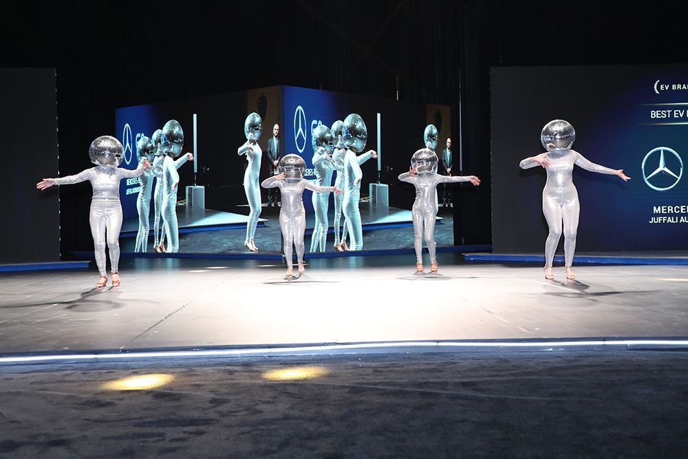 Performers in silver suits and helmets dance on stage in front of a screen.