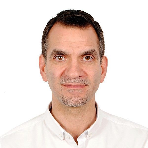 A man with short brown hair, stubble, wearing a white shirt, and looking forward with a slight smile.