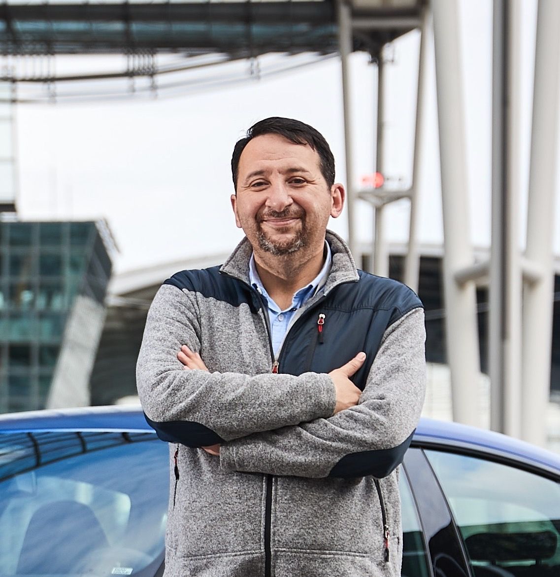 Man in gray jacket with arms crossed, smiles near blue car. Outdoors with modern building.