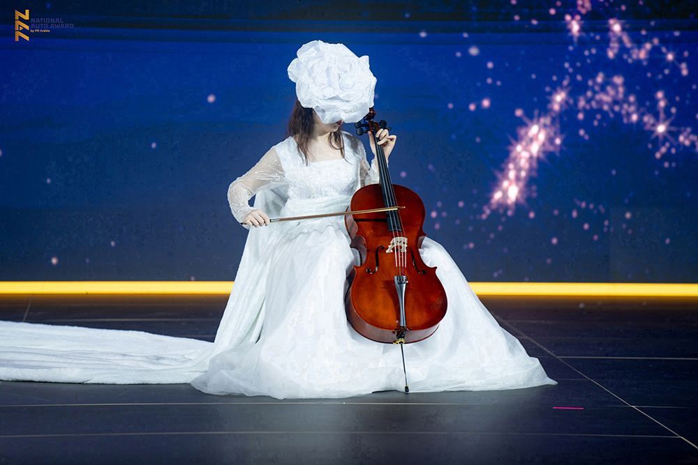 Woman in white gown and floral face mask playing a cello on stage; sparkly background.