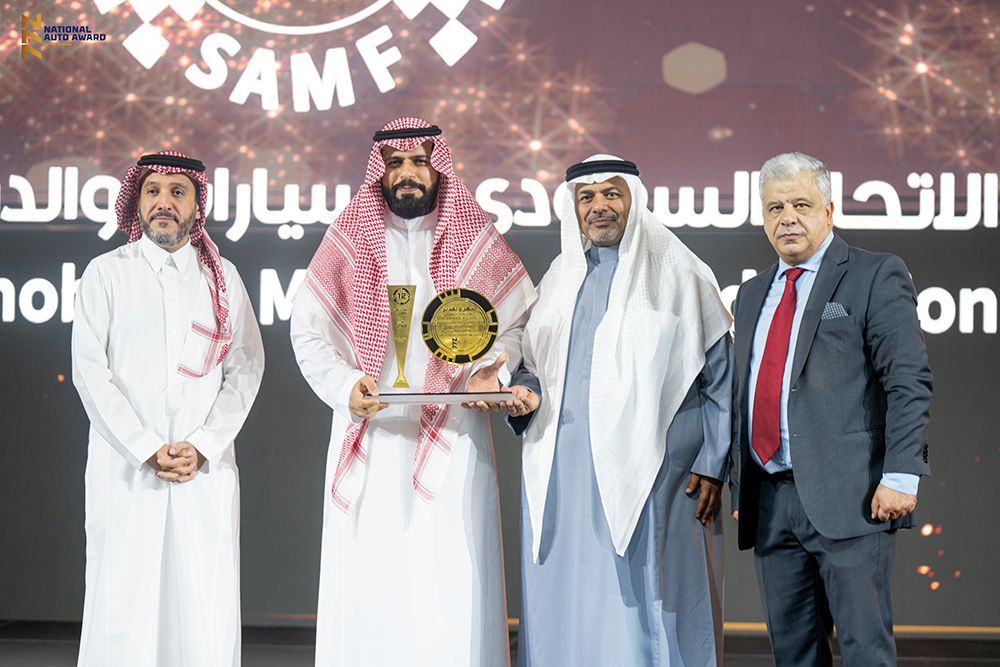 Four men on stage, one holding a trophy. They are in traditional Arab dress with a dark backdrop, lights and text.