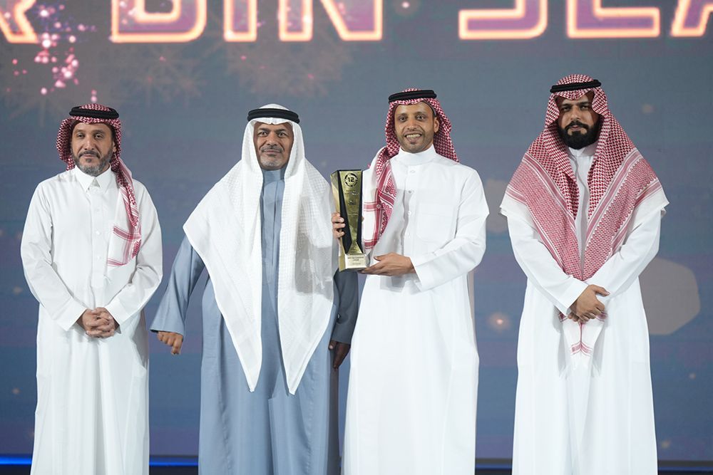 Four men in white thobes and ghutras on stage, one holding a trophy. Dark background.