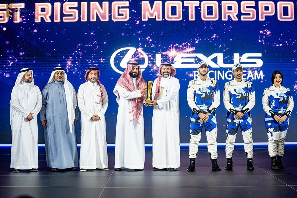 Group of people in traditional white robes and team members in blue uniforms hold a trophy. Lexus Racing Team.