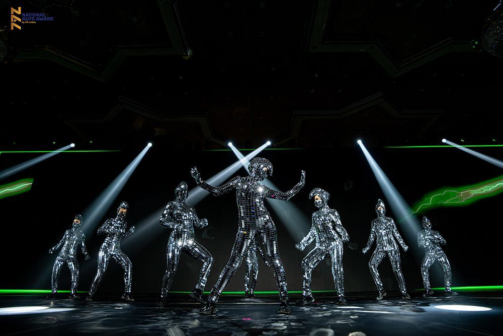 Seven figures in silver metallic suits dance under spotlights on a green-lit stage.
