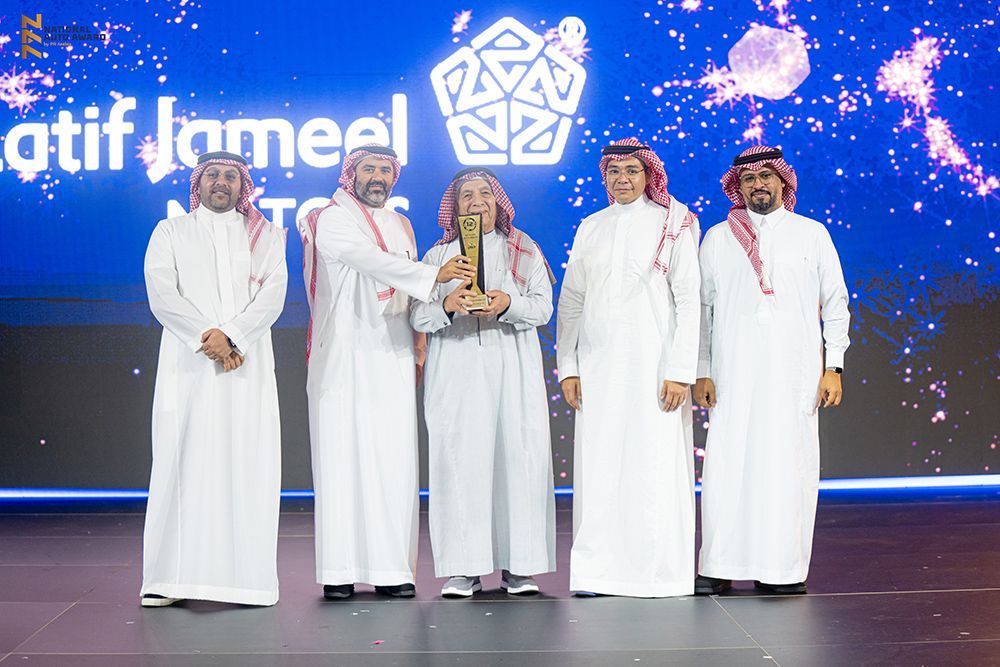 Five men in white thobes on stage; one receives an award. Blue background with a logo, glitter.
