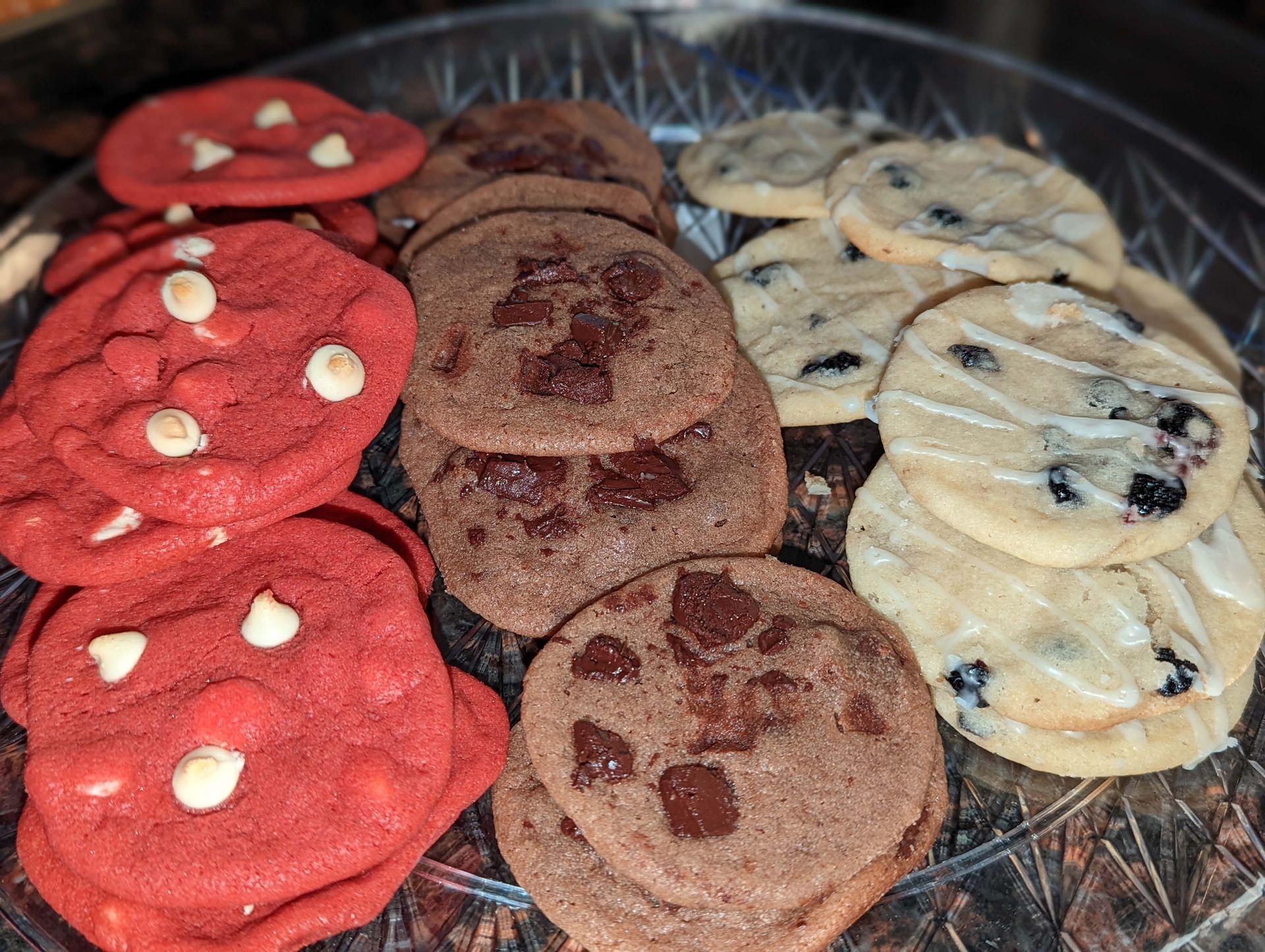 A glass plate topped with a variety of cookies including red velvet cookies , chocolate chip cookies , and blueberry cookies.