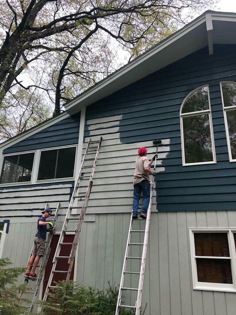 A man is standing on a ladder painting the side of a house.